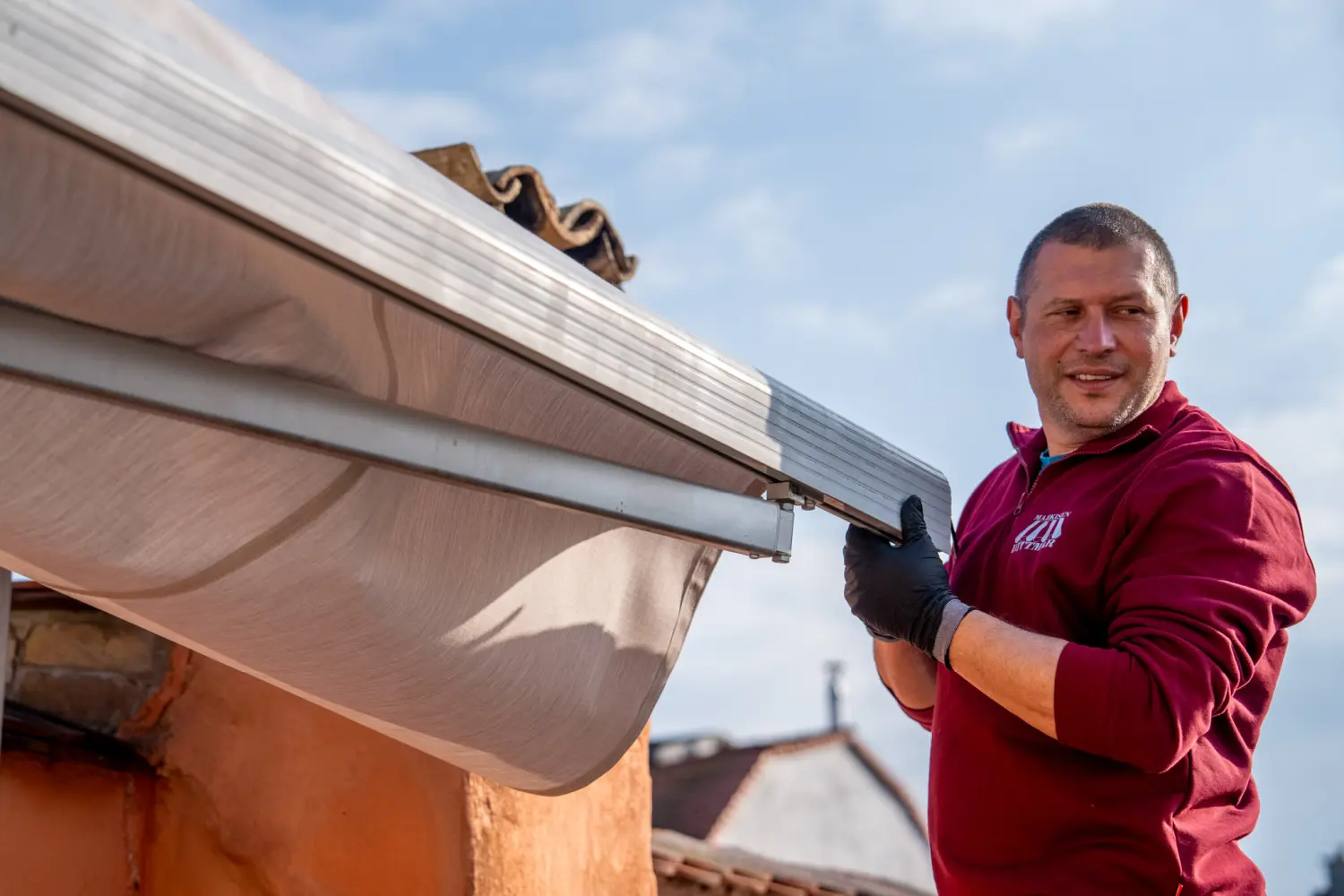 Eine Person im roten Hemd bewältigt gekonnt die Montage einer Markise auf dem Gebäude vor einem klaren blauen Himmel und ist ein Beispiel für die wahre Fachbetriebskompetenz.