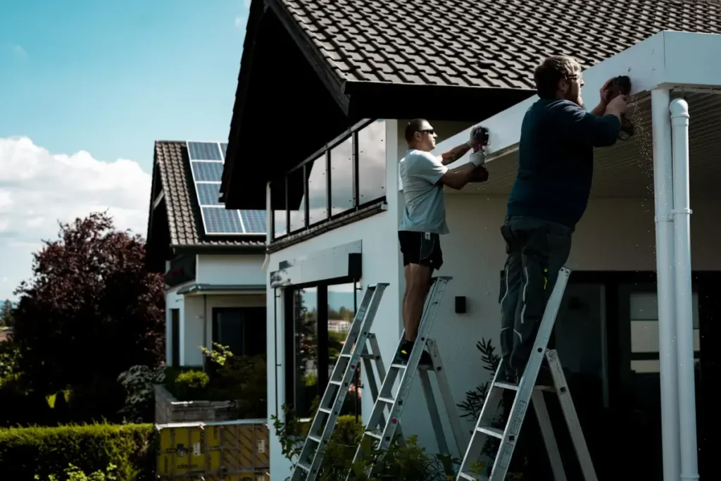 Zwei Männer auf Leitern befestigen eine weiße Dachrinne auf einem Hausdach mit Solarmodulen, umgeben von Grün.