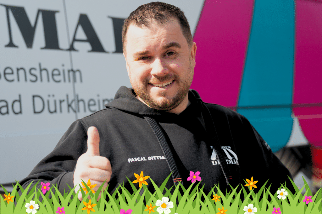 Ein lächelnder Mann zeigt den Daumen nach oben und steht im Freien mit Blumen und Gras im Vordergrund.