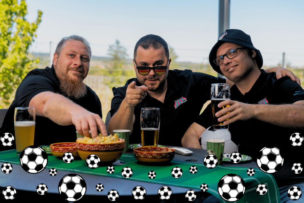 Drei Männer an einem Tisch mit Snacks und Bier, lächelnd, mit einer Fußballgrafik am unteren Rand des Bildes.